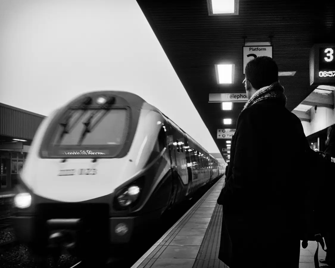 Schwarzweißaufnahme: eine Person wartet am Bahnsteig, ein Triebzug fährt ein