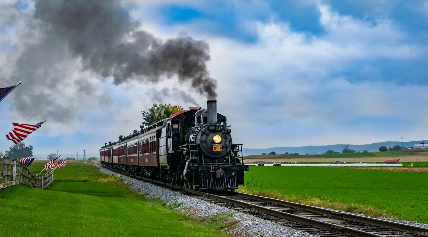 Schwarze Dampflokomotive mit Wagenzug auf freier Strecke, weiße Dampfwolke über dem Kessel