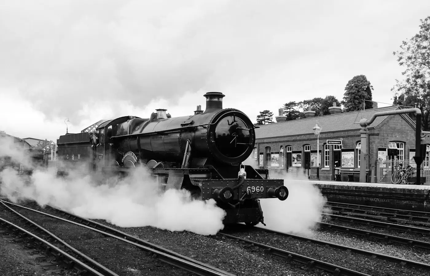 Historische Schwarzweißaufnahme einer Dampflokomotive, die vor einem Bahnhofsgebäude abdampft
