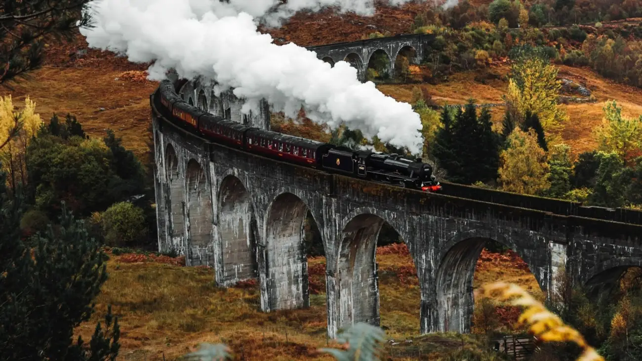 Dampfzug mit langer Rauchfahne auf einem gemauerten Bogenviadukt in hügeliger Landschaft