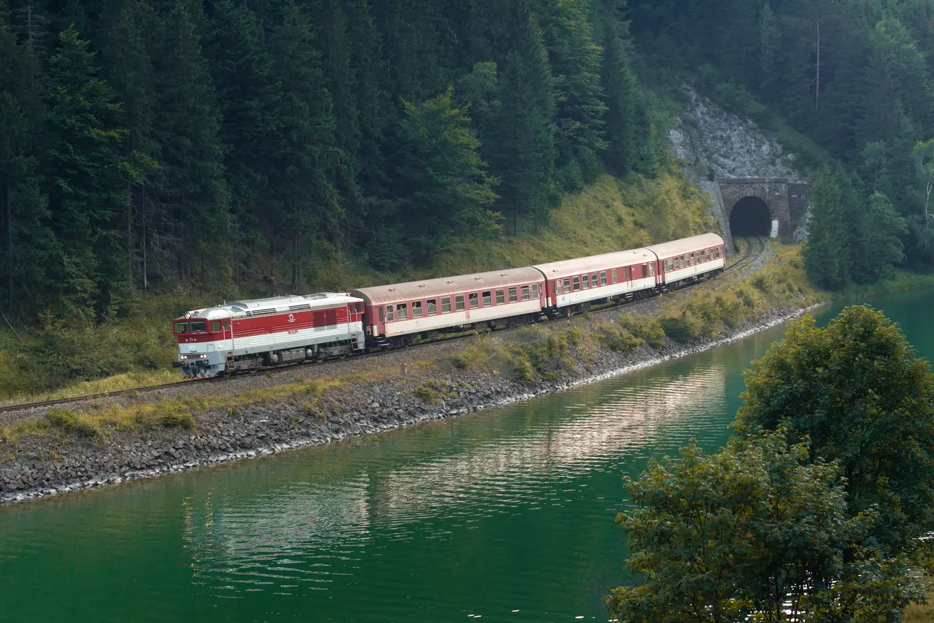 Ein rot-weißer Reisezug fährt am Ufer eines türkisfarbenen Sees auf einen Tunnel zu